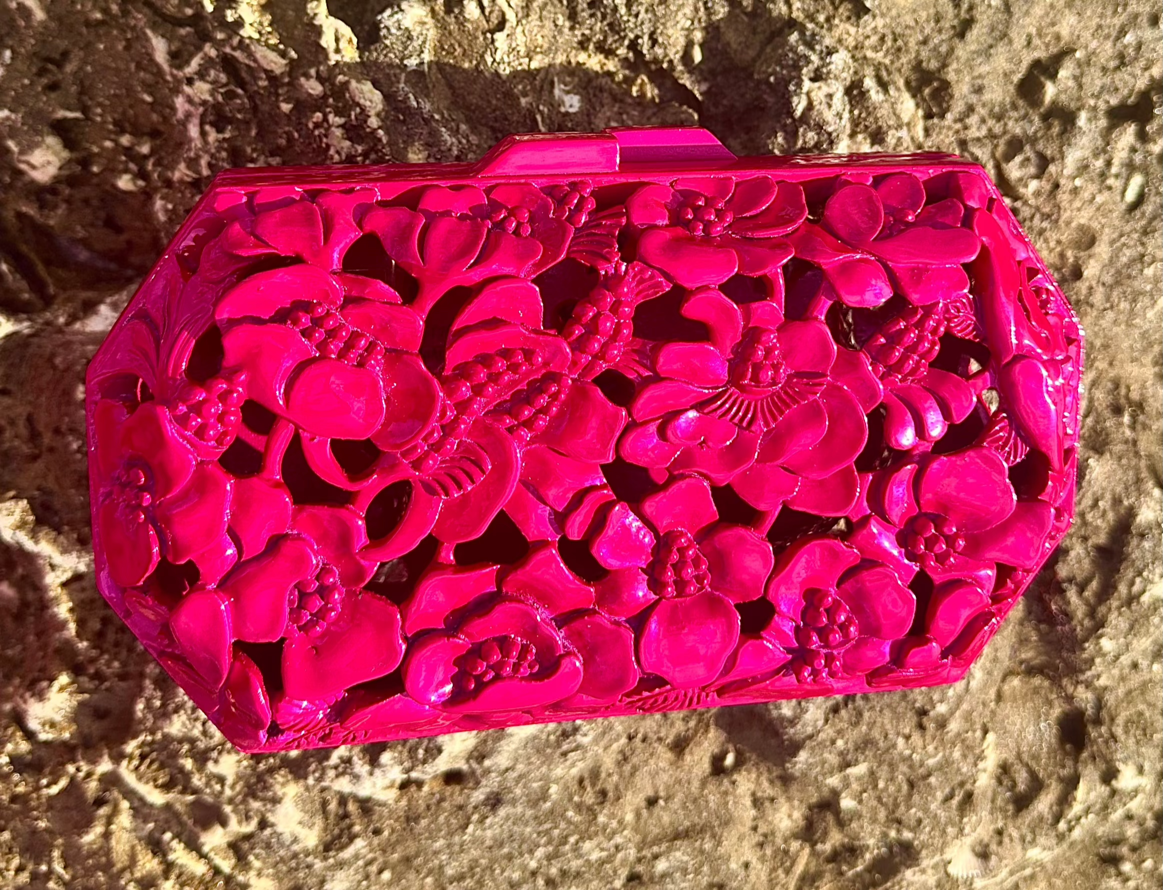 Hand-carved magenta teak wood clutch by Blumera resting on a natural rock, radiating vibrant color and sculptural elegance. A collectors favorite.