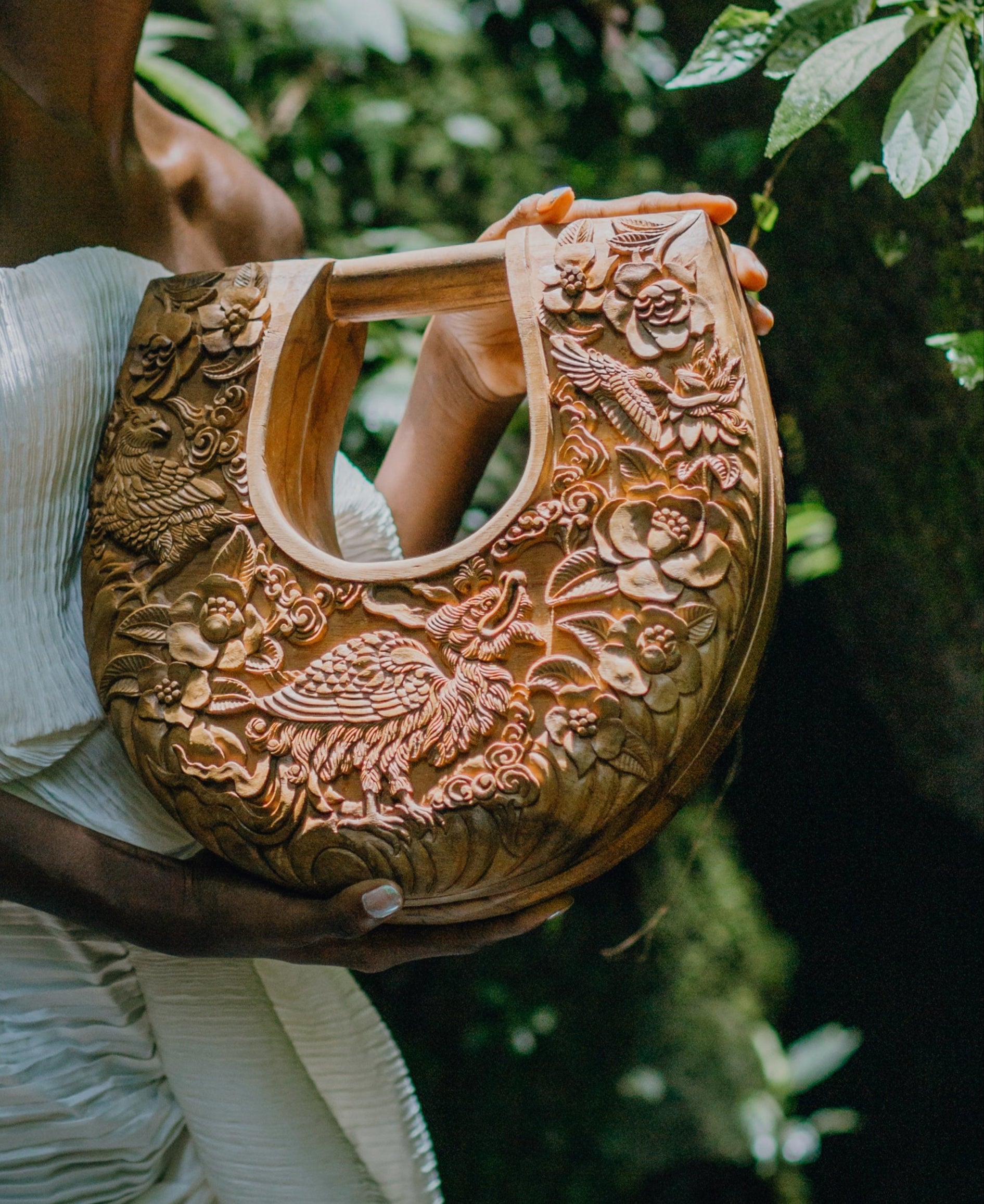 Intricately hand-carved teak wood handbag by Blumera held by model in a serene moss setting, inspired by Laurie Blum’s birds and floral paintings.